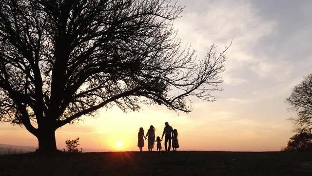 Silhouettes of happy family walking in the meadow near a big tree during sunset.
