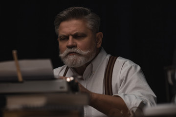 serious senior writer working with typewriter isolated on black