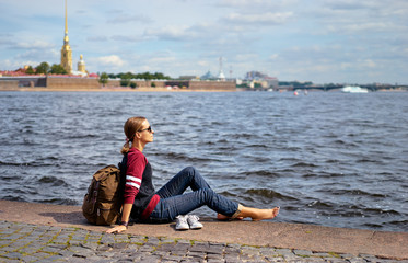 Travel and tourism. Young woman sitting on embankment of Neva river enjoying the view of Peter and Paul fortress, Saint Petersburg, Russia.