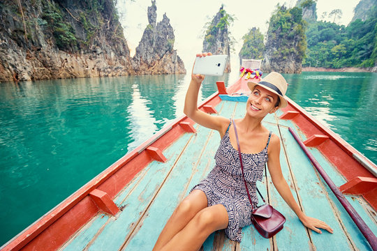Happy vacation in Thailand. Pretty young woman taking selfie on smartphone sailing Khao Sok National Park lake on traditional longtail boat.