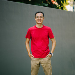 A portrait of a cool, middle aged Asian man standing against the gray wall during the day. He is looking casual on his red tee and khaki pants as he looks so decent and calm.