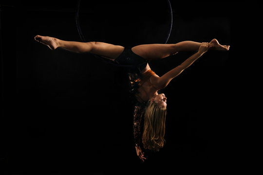 Young female aerial acrobat doing splits hanging upside down from hoop, black background