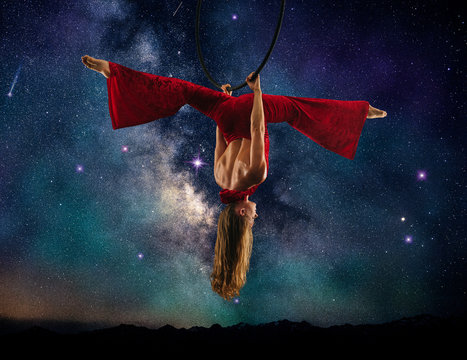 Young female aerial acrobat doing splits hanging upside down from hoop, milky way background