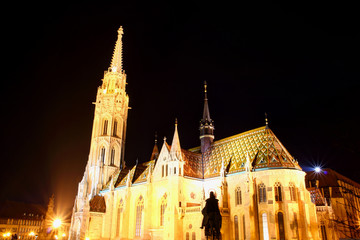 Fototapeta premium Low angle view of majestic tall Matthias Church floodlit by exterior facade lights with King Stephen equestrian statue silhouette Buda Castle Fisherman's Bastion Trinity square Budapest Hungary Europe