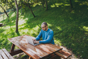 Fototapeta premium Young successful smart man businessman or student in casual blue shirt glasses sitting at table with mobile phone in city park using laptop working outdoors on green nature. Mobile Office concept.