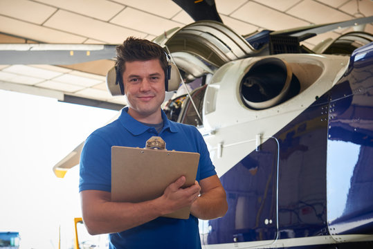 Portrait Of Male Aero Engineer With Clipboard Carrying Out Check On Helicopter In Hangar