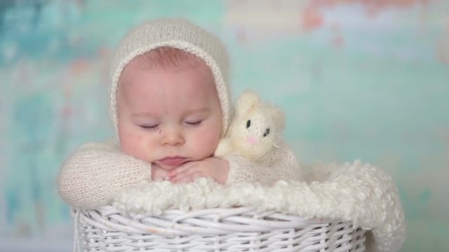Little cute baby boy, dressed in handmade knitted white teddy bear overall, sleeping peaefully with little knitted toy in basket