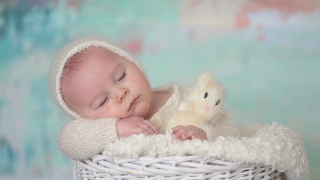 Little cute baby boy, dressed in handmade knitted white teddy bear overall, sleeping peaefully with little knitted toy in basket