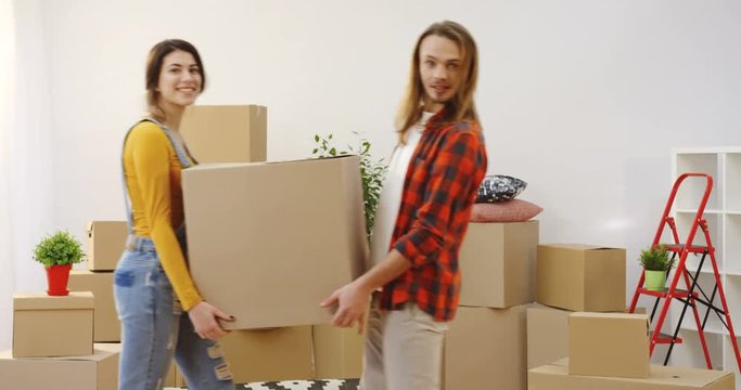 Young cute attractive couple having fun while moving in the new apartment,they carrying abox and plant in front of the camera. Boxes on the background. Indoor