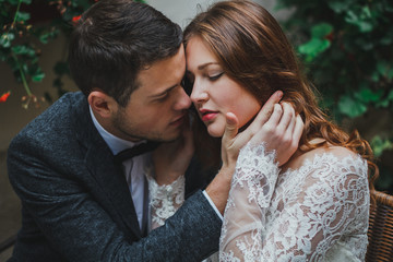 Wedding couple hugs in old city vintage street cafe. He takes her face in hands in ancient town. Rustic bride in lace tulle dress white with hair down and groom in grey suit and bow tie. Stone walls.