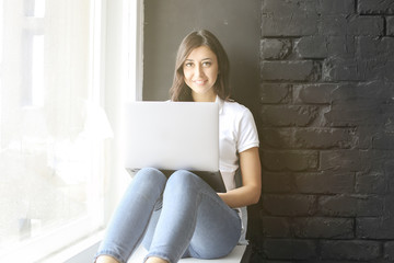 Happy millennial girl w/ laptop on windowsill. Portrait of young woman with diastema gap between teeth. Beautiful smile. Minimal interior, black brick textured wall background, loft style. Copy space.