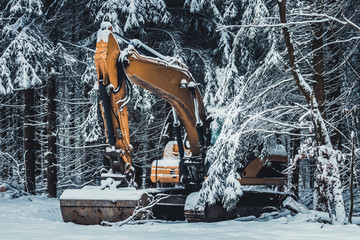 Yellow excavator heavy industry machine standing under the trees in a dark snowy forest