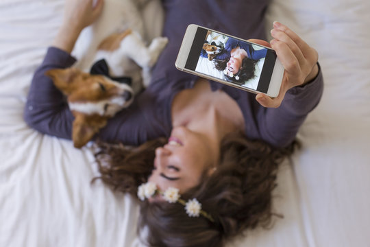 Top View Of A Beautiful Young Woman Taking A Selfie With Mobile Phone On Bed With Her Cute Small Dog Besides. Home, Indoors And Lifestyle