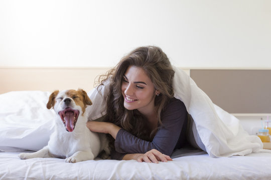 beautiful young woman lying on bed with her cute small dog besides and yawning. Home, indoors and lifestyle