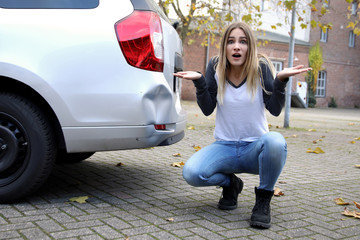 Hübsche blonde Frau neben einem Auto mit Unfallschaden ratlos