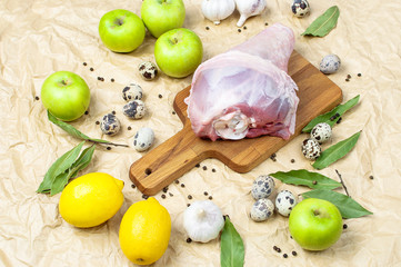 Raw turkey thigh garlic apples lemon quail eggs spices on a wooden board. Top view of a turkey, knife on a beige paper background. Culinary background, ingredients for cooking.