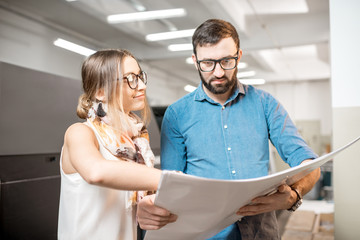 Young woman designer and print operator working together with paper print standing at the print manufacturing