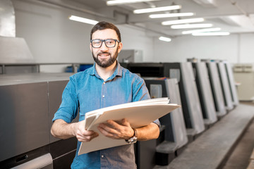 Portrait of a handsome typographer standing with stack of paper at the printing manufacturing with...
