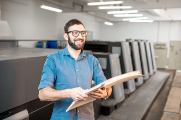 Portrait of a handsome typographer standing with stack of paper at the printing manufacturing with...
