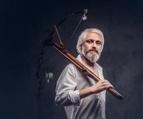 Studio portrait of a smiling handsome old man with a gray beard and white shirt holding a crossbow on shoulder.