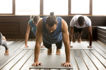 Group of young sporty people practicing yoga lesson standing in Plank pose, doing Push ups or press...