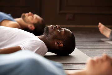 Group of young afro american and caucasian sporty people practicing yoga lesson lying in Dead Body or Corpse pose, Savasana exercise, resting after practice, working out, indoor close up, studio - Powered by Adobe