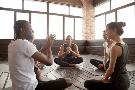 Excited african american man talking to sporty happy diverse friends sitting on mat in studio, happy black yoga instructor or life coach motivating young people having fun at seminar training class
