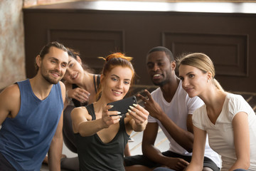 Attractive sporty smiling woman holding phone taking group selfie at training class in gym studio, diverse fit people, happy fitness friends or multiracial yoga team make self portrait on smartphone