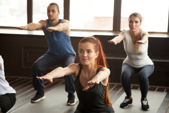 Sporty woman doing squat exercise at fitness training with young people, fit motivated group warming up muscles at strength training class working out together during routine session in gym studio