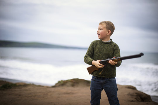 Young boy standing on a beach while holding a rifle.