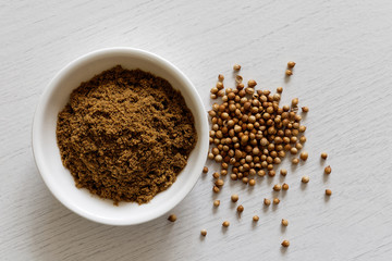 Ground coriander seeds in white ceramic bowl isolated on white wood background from above. Whole dried coriander seeds.