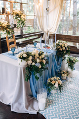 Table with blue tablecloth decorated with flowers and green branches, candles in tall vases next to table, light bulb garland on background