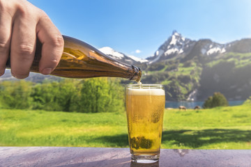 Glass of beer and summer alpine landscape - Hand of a man pouring beer from a bottle in a glass with a summer scenery in the background with mountains and green meadows, on a sunny day. © YesPhotographers