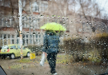 Car windshield with rain drops during falling rain and blurred man with umbrella. Shallow depth of field with focus on center of the windshield and urban background