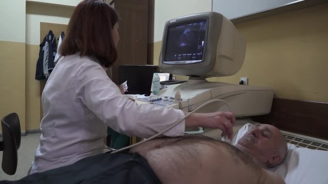 Cardiology examination. Female doctor cardiologist using ultrasound device, looking in computer monitor and talking with male patient who lying on bed in room, nurse writing in background, dolly shot