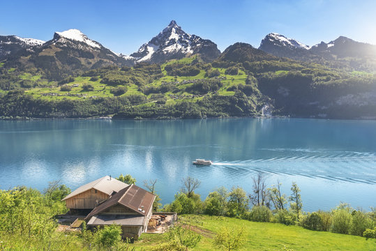 Swiss alps and mountain lake in spring - Amazing spring landscape with the Alps mountains, the Walensee lake with a boat floating on it, and an old stable on a green meadow, on a sunny day.