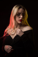 Beautiful blonde woman in glasses wearing blouse with naked shoulders, posing with red and yellow light