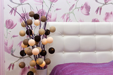 Cotton light balls string in brown, beige and white colors in beautiful pink bedroom interior with stylish bed and decorative vase.