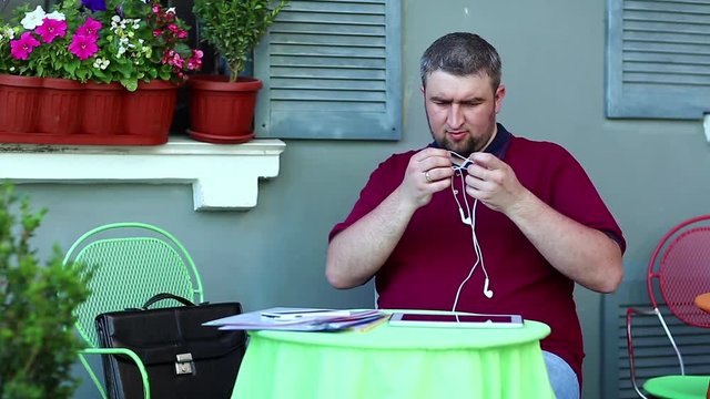 Businessman sits at the table in summer restaurant and disentangle earpieces for tablet computer. Man untangle earphones