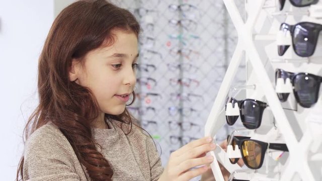 Close up of a little pretty girl looking excited examining sunglasses for sale at the eyewear optometrist store shopping buying consumerism fashion style vision optics kids children leisure.