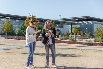 Two women with black glasses check their mobile phones, in the middle of a park
