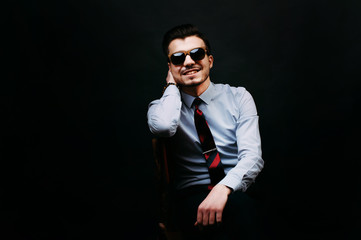 Fototapeta premium Handsome smiling and cheerful young business man in casual sitting on chair and wearing sunglasses and looking at the camera over dark background.