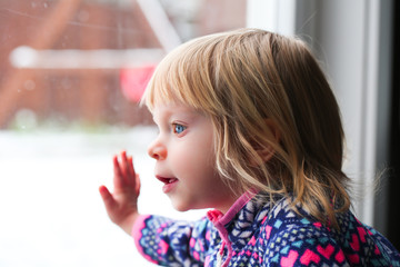 Close-up Portrait of Cute Blonde 18 Month Old Toddler Girl with Big Blue Eyes, looks at the window, Sweet Face Expression, Stand by the Window, Snowy Background 