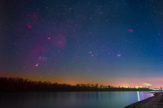 Winter night sky with Barnard’s Loop, the Orion Nebula, the Flame Nebula, the Rosette Nebula, the California Nebula, and the Pleiades as seen from the shore of the river Rhine at Mannheim in Germany.