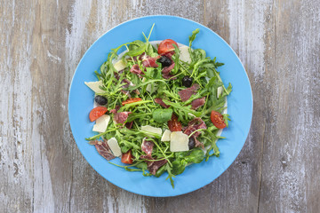 Fresh salad - Delicious fresh salad with tomatoes, lettuce, eggplant, zucchini, cheese, parma ham and olive oil, blue plate, grey wooden background