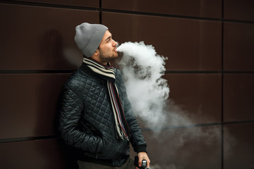 Vape man. Portrait of a handsome young white guy in casual clothes vaping an electronic cigarette and letting out puffs of steam opposite the futuristic modern building. Lifestyle. Close up.