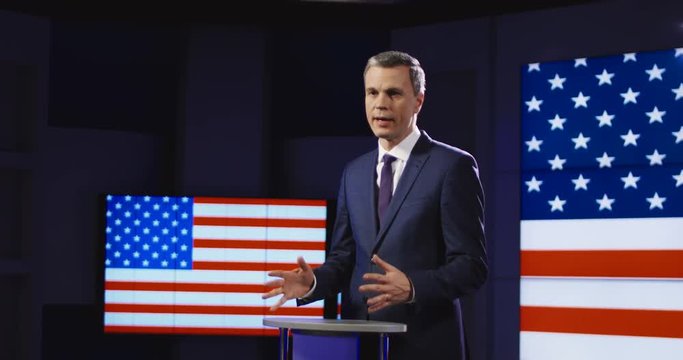 Public speaker or presenter in front of American flag standing at a small rostrum speaking and gesturing emphatically with his hands. 4K shot on Red cinema camera.