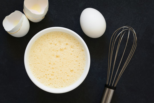 Whisked whole eggs (whites and yolks together) in bowl, with whisk and egg shells on the side, photographed overhead on slate with natural light