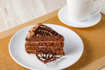 The slice of chocolate cake Prague with chocolate buttercreamy frosting on a white plate. Cup of coffee near it. Homemade baking. Close-up view.