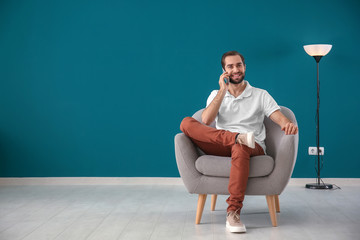 Handsome man talking on mobile phone while sitting in comfortable armchair against color wall